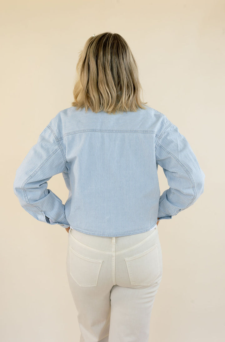 A woman viewed from behind wearing a light blue striped cropped jacket and cream-colored pants, standing against a solid beige background.