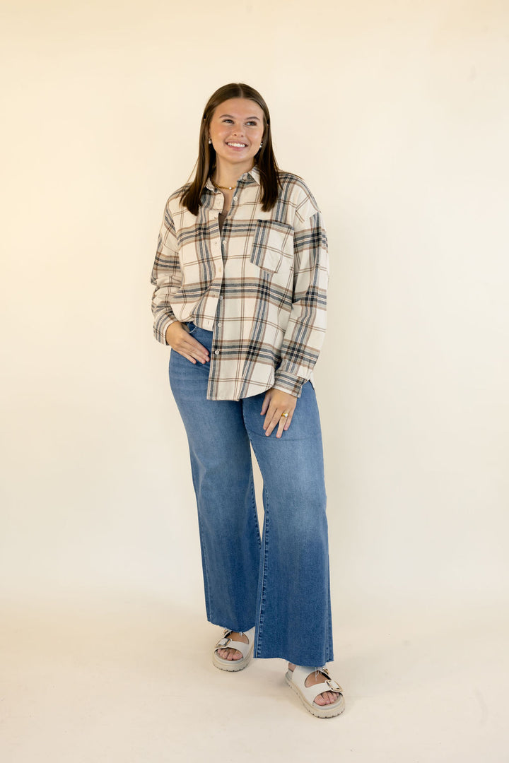 A woman modeling a long-sleeve plaid button-up shirt in beige, brown, and navy, paired with wide-leg blue jeans and white slide sandals, standing against a plain cream studio background.