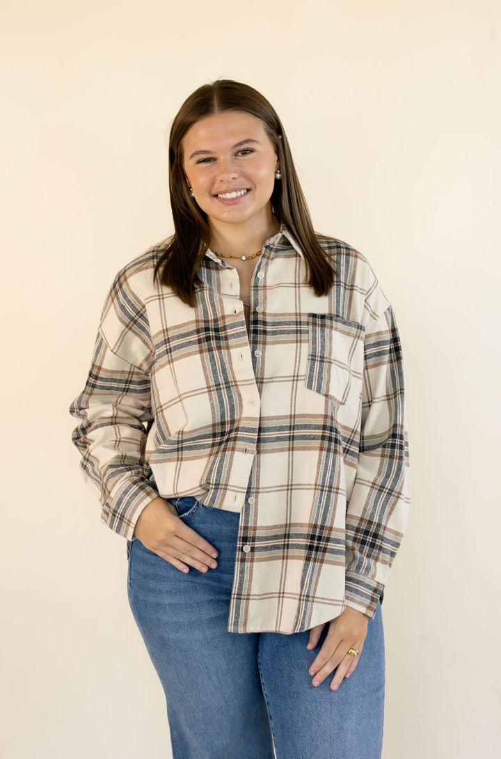A woman modeling a long-sleeve plaid button-up shirt in beige, brown, and navy, paired with blue jeans, against a plain cream background.
