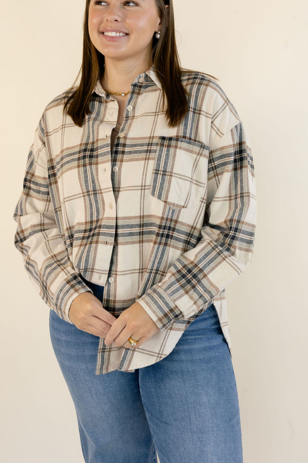 A woman wearing a long-sleeve button-up plaid shirt in beige, brown, and navy, paired with blue jeans, standing against a plain off-white background.