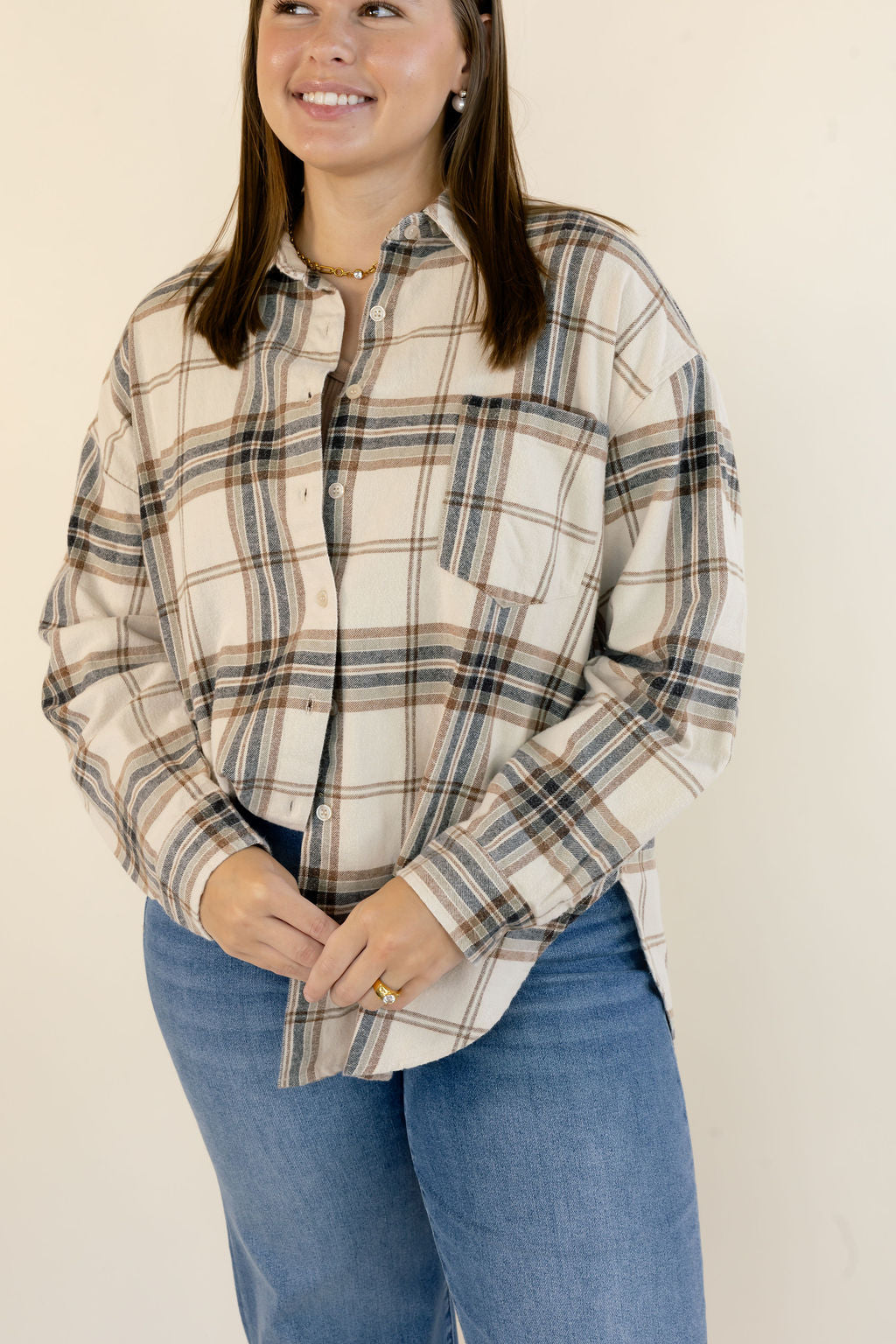 A woman wearing a long-sleeve button-up plaid shirt in beige, brown, and navy, paired with blue jeans, standing against a plain off-white background.