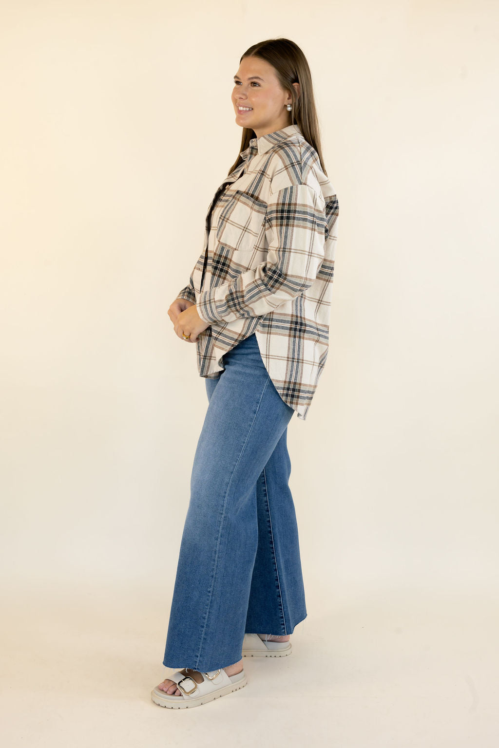 A woman modeling a long-sleeve plaid button-up shirt in cream, beige, and navy, paired with wide-leg blue jeans and white slide sandals, standing against a plain off-white studio background.