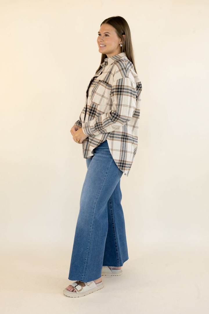 A woman modeling a long-sleeve plaid button-up shirt in cream, beige, and navy, paired with wide-leg blue jeans and white slide sandals, standing against a plain off-white studio background.