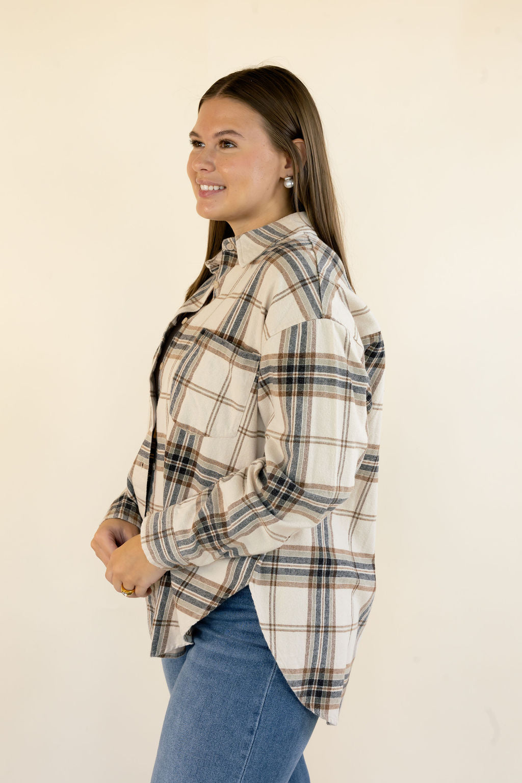 A woman wearing a cream-colored plaid button-up shirt with blue jeans, standing against a plain off-white background.