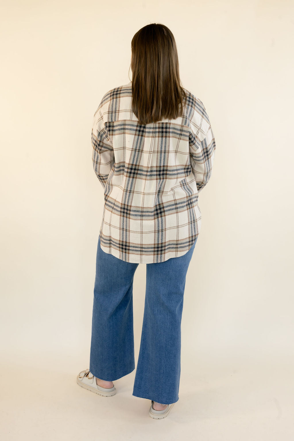A woman viewed from behind wearing an oversized cream and brown  and navy plaid button-up shirt and wide-leg blue jeans, standing against a plain off-white studio background.