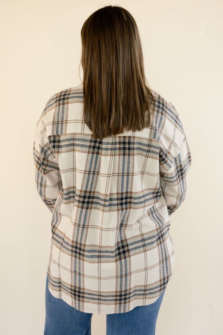 A woman viewed from behind wearing an oversized plaid flannel shirt in cream, brown, and navy, paired with blue jeans, against a plain off-white background.