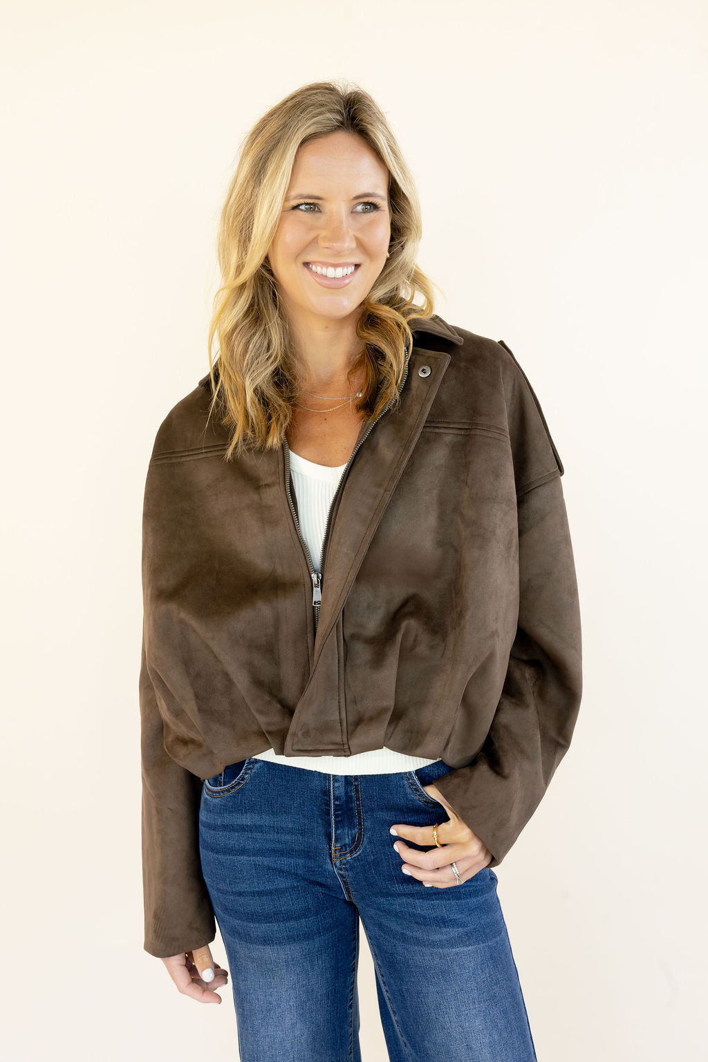 Woman modeling dark brown faux suede cropped jacket with zip front, collared neckline, epaulet details, and blue flare jeans