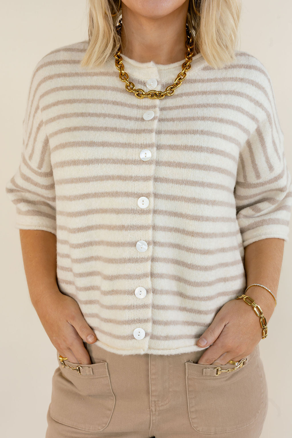 A woman wearing a cream and taupe striped knit cardigan with white buttons, styled with beige pants and gold jewelry against a light beige background.