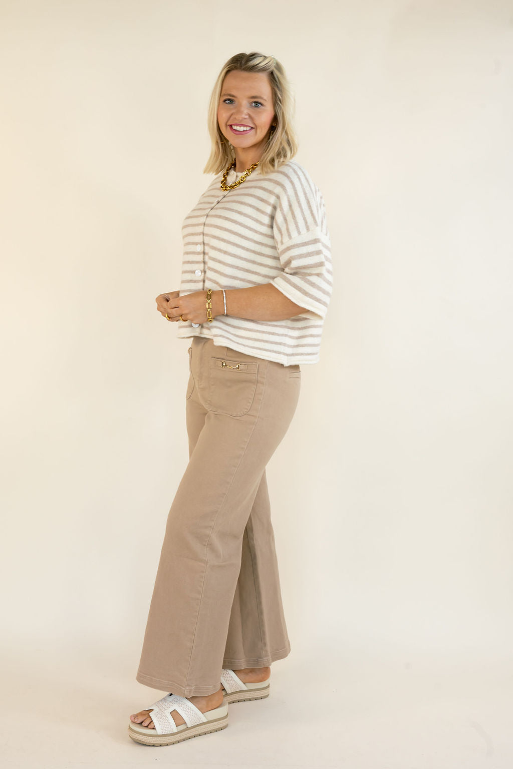 A woman modeling a cream and tan horizontally striped knit top with elbow-length sleeves, paired with beige wide-leg pants and white slide sandals, standing against a plain cream studio background.