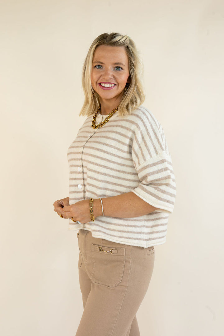 A smiling blonde woman wearing a beige and white striped knit cardigan with three-quarter sleeves, paired with beige pants, standing against a plain cream background.