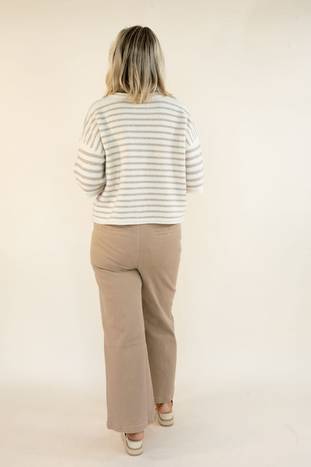 A woman viewed from behind wearing a cream and tan striped long-sleeve knit top and beige wide-leg pants, standing against a plain off-white studio background.