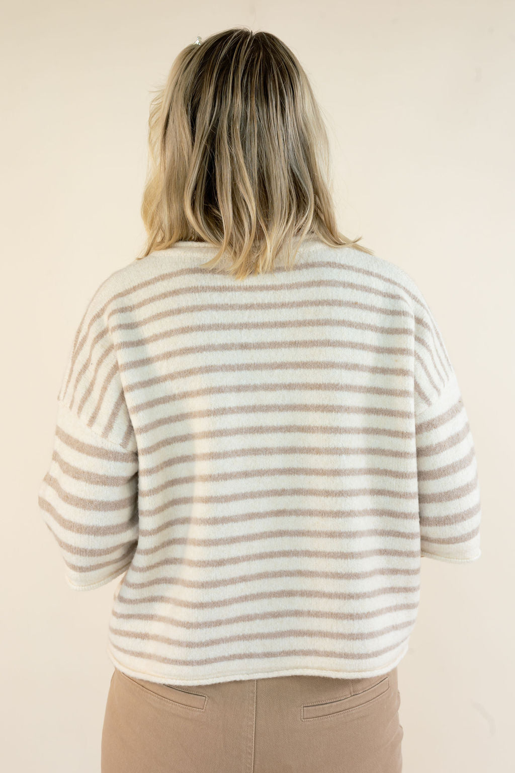 A woman viewed from behind wearing a fuzzy knit sweater with horizontal beige and off-white stripes, paired with tan pants, against a plain cream background.
