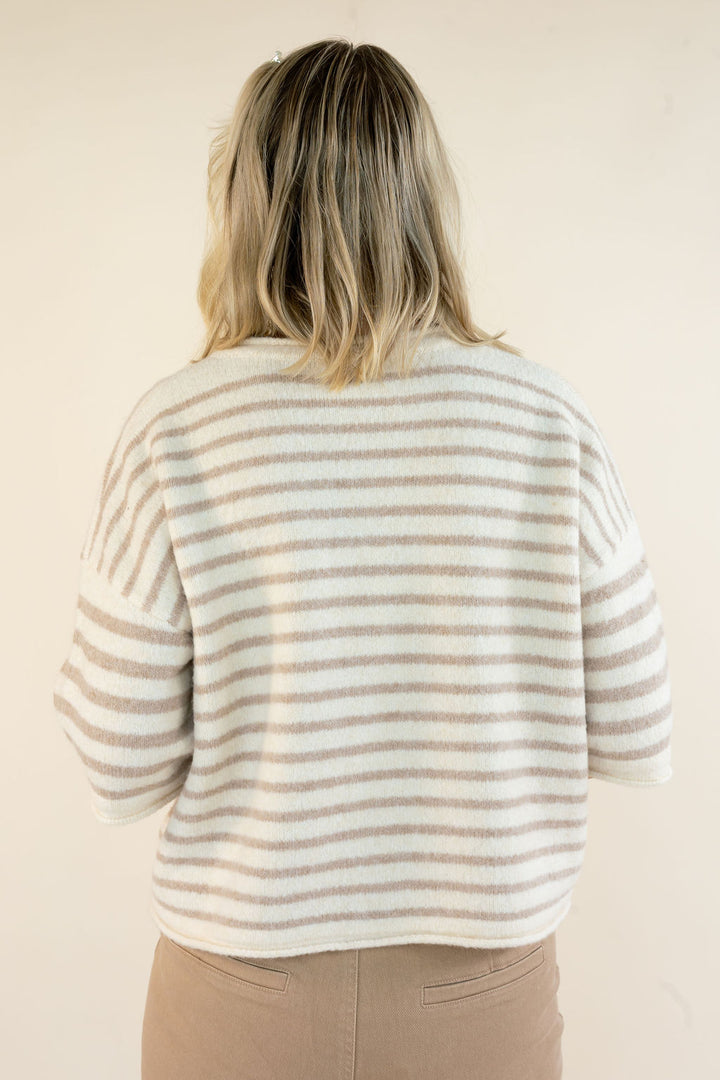 A woman viewed from behind wearing a fuzzy knit sweater with horizontal beige and off-white stripes, paired with tan pants, against a plain cream background.