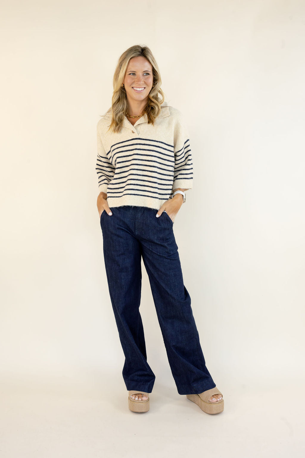 A woman modeling a cream and navy blue horizontally striped knit polo sweater paired with dark blue wide-leg jeans and beige platform sandals, standing against a plain off-white studio background.
