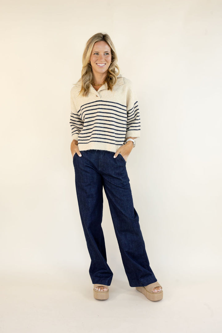 A woman modeling a cream and navy blue horizontally striped knit polo sweater paired with dark blue wide-leg jeans and beige platform sandals, standing against a plain off-white studio background.