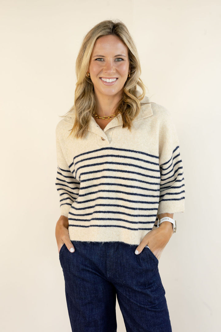 A smiling blonde woman wearing a cream knit polo sweater with navy horizontal stripes and dark blue jeans, standing against a plain off-white background.