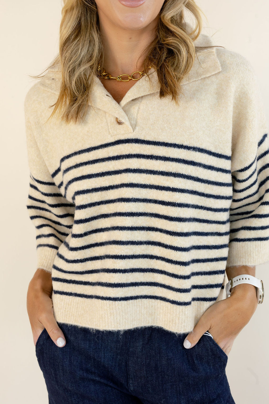 A woman wearing a cream knit polo sweater with navy horizontal stripes and dark blue jeans, photographed against a plain light beige background.