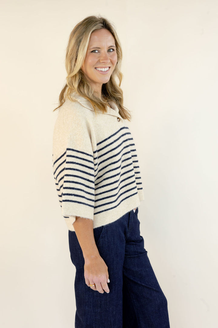 A woman modeling a cream knit cropped sweater with navy horizontal stripes on the sleeves and upper chest, paired with dark blue wide-leg pants, against a plain off-white background.
