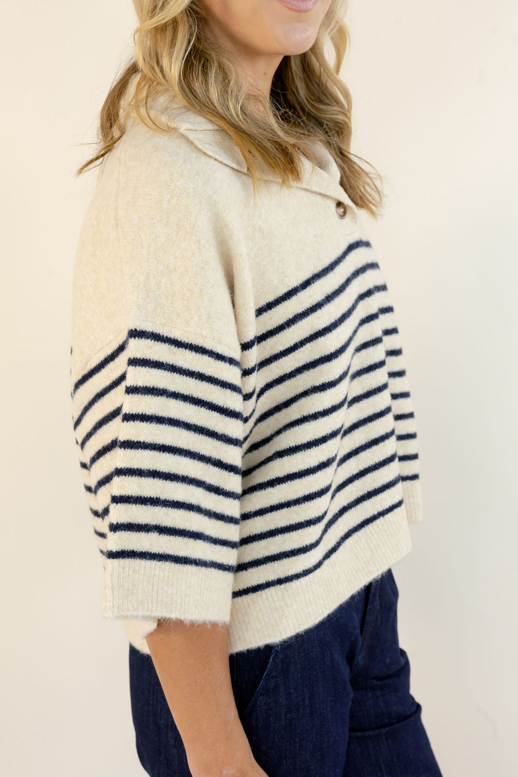 A woman wearing a cream knit cropped sweater with navy horizontal stripes on the sleeves and upper chest, paired with dark blue jeans, photographed against a plain light beige background.