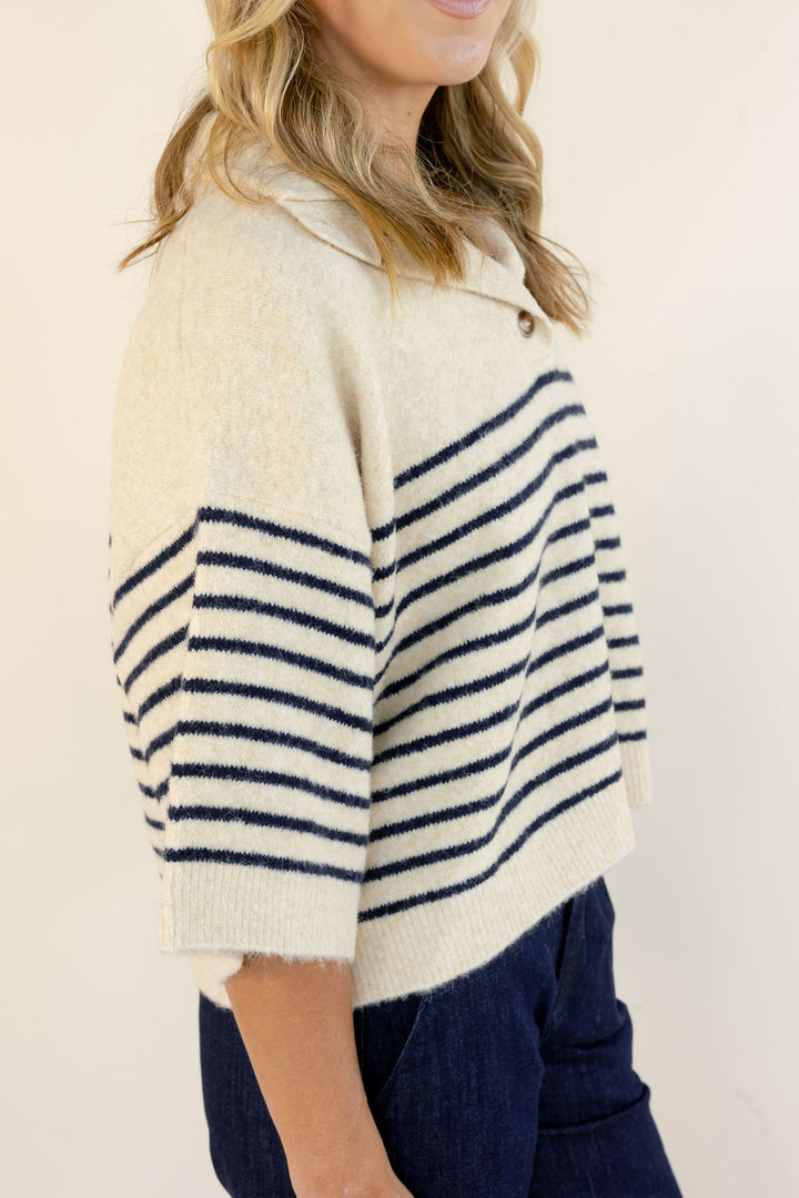 A woman wearing a cream knit cropped sweater with navy horizontal stripes on the sleeves and upper chest, paired with dark blue jeans, photographed against a plain light beige background.
