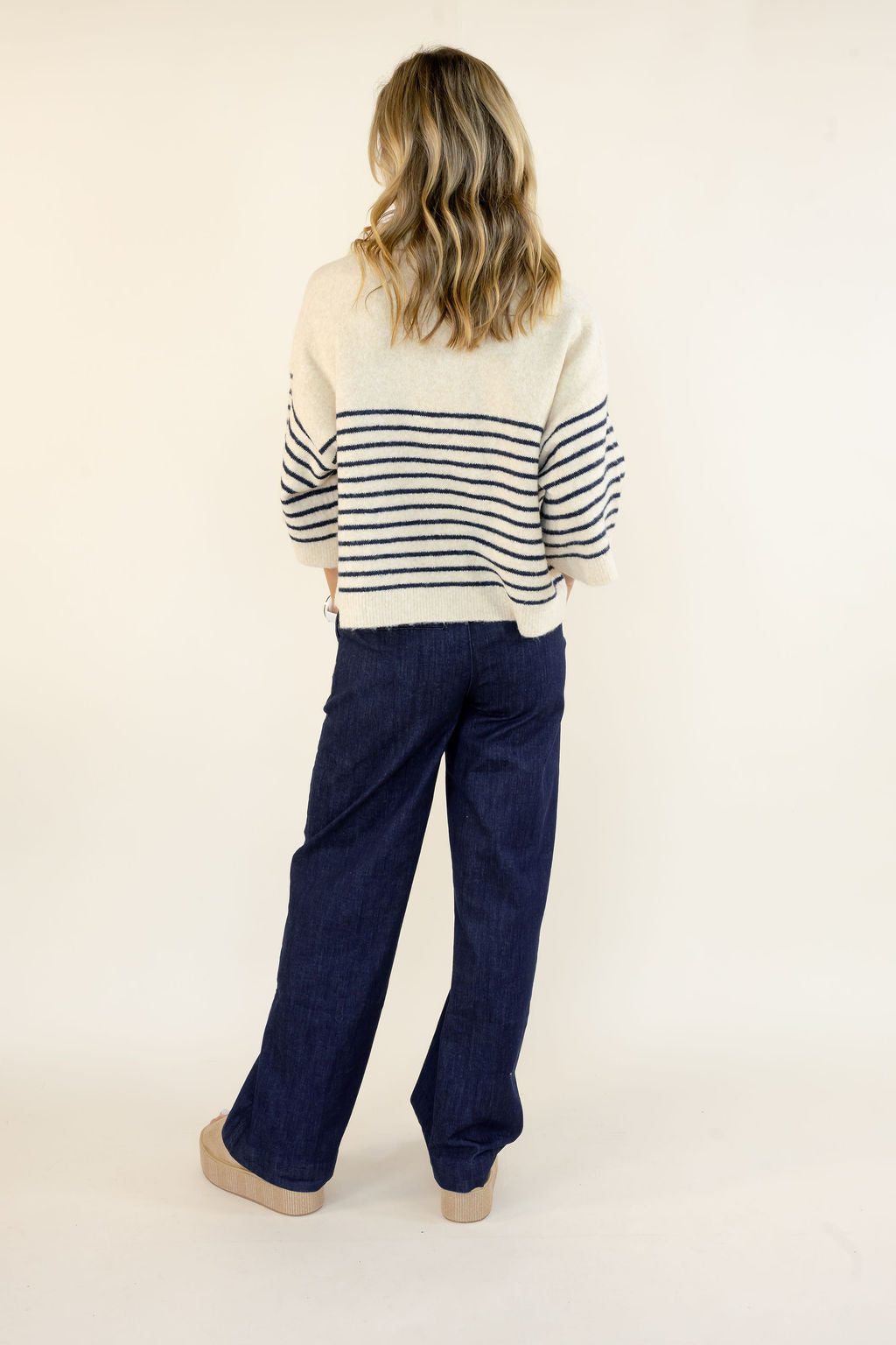 A woman shown from behind wearing a cream knit sweater with navy horizontal stripes on the sleeves and upper back, paired with dark blue wide-leg pants and beige shoes, against a plain off-white studio background.