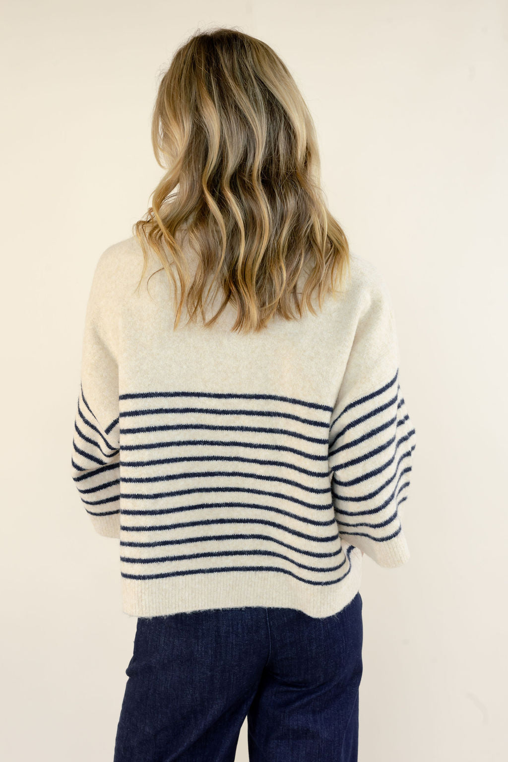 A woman viewed from behind wearing a cream knit sweater with navy horizontal stripes across the back and sleeves, paired with dark blue jeans, against a plain off-white background.