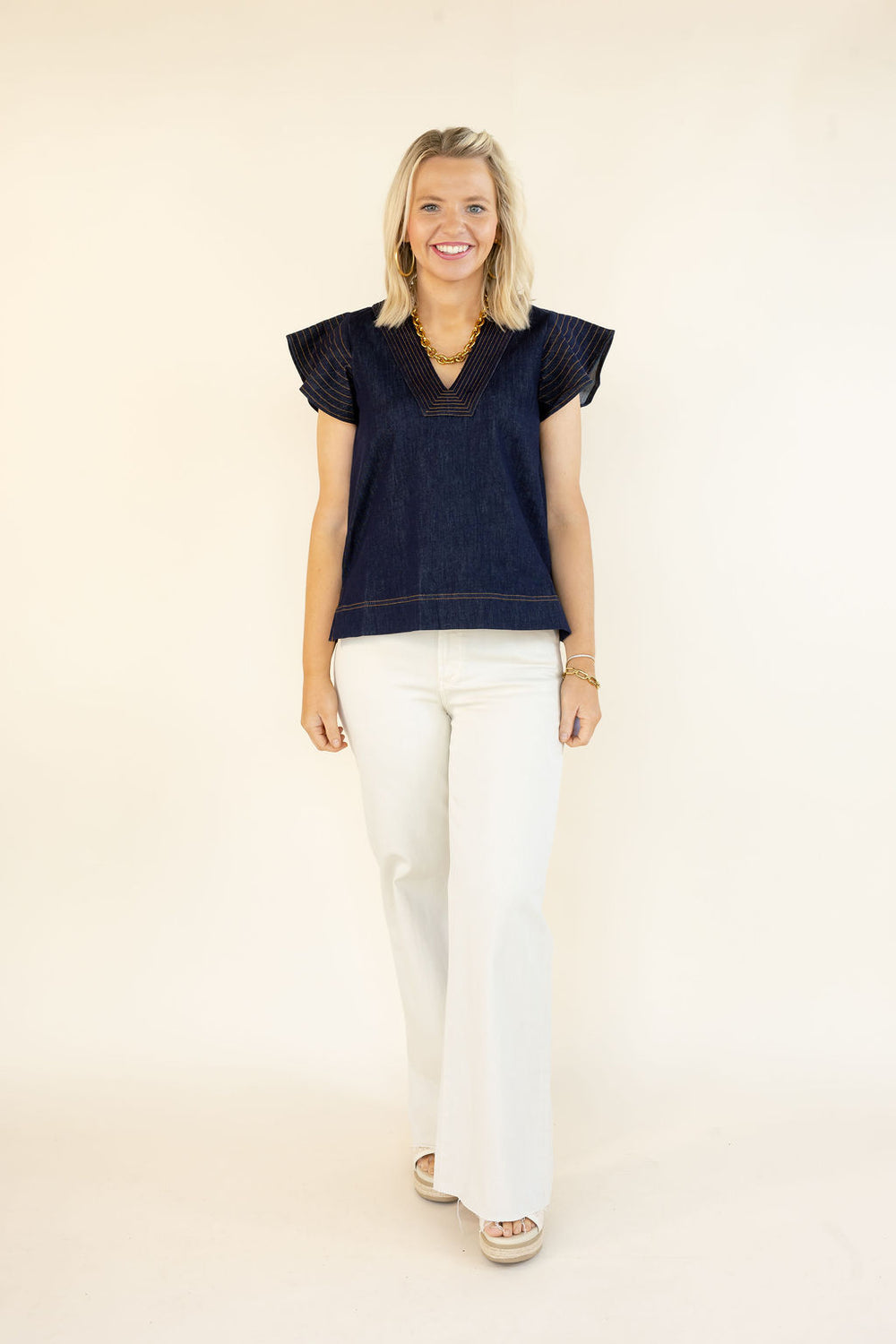 Woman modeling THML denim blue V-neck top with layered flutter sleeves, contrast stitching, and white flare jeans
