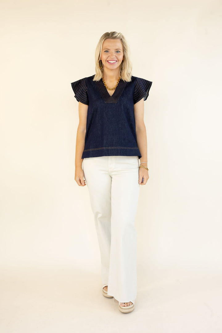 Woman modeling THML denim blue V-neck top with layered flutter sleeves, contrast stitching, and white flare jeans