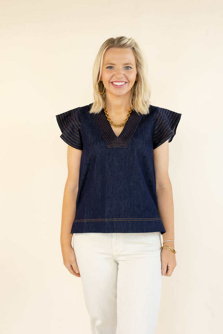Front view of THML denim blue short sleeve top featuring a V-neckline, statement flutter sleeves, and relaxed fit styled with white jeans