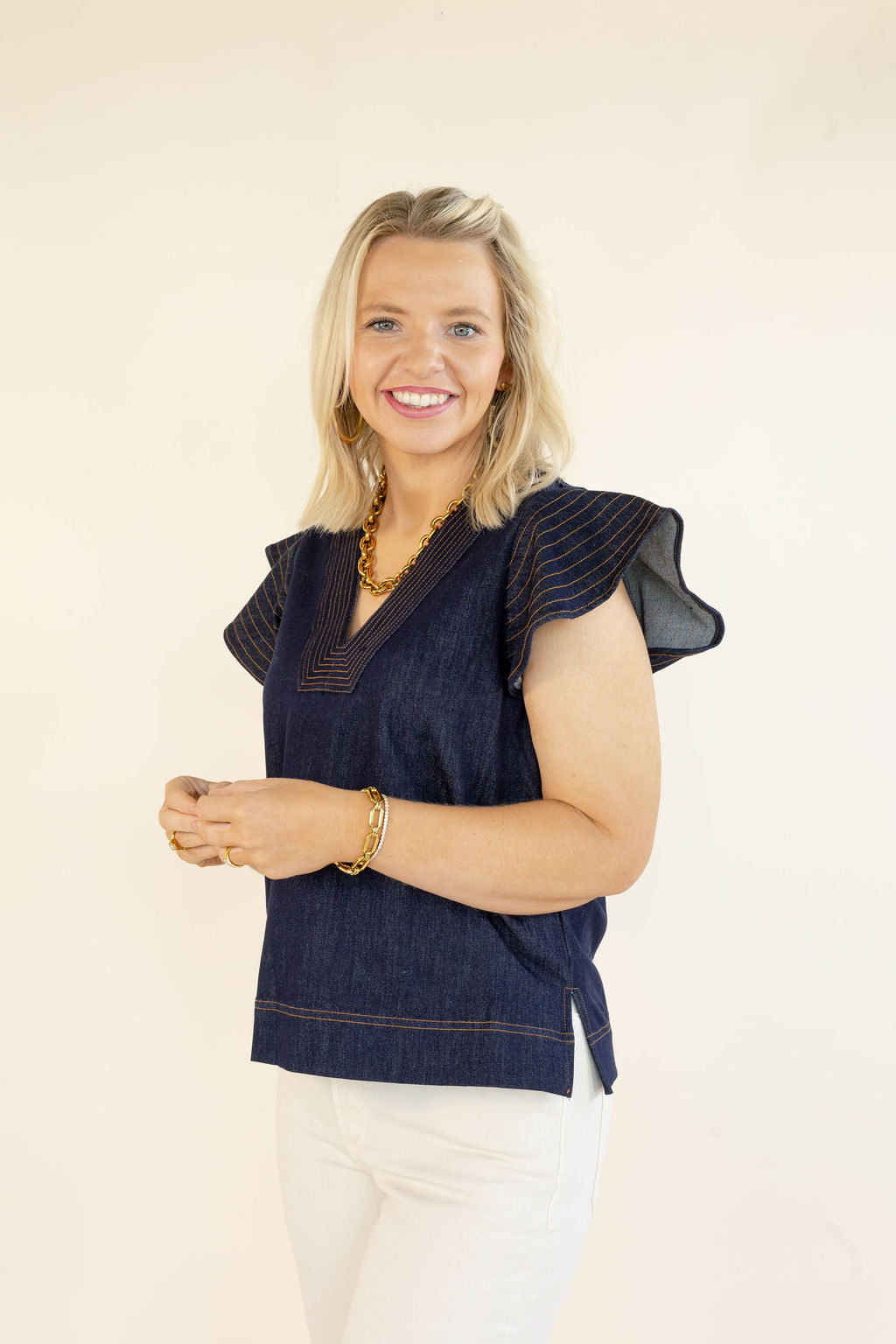Three-quarter view of woman wearing THML denim blue V-neck top with layered flutter sleeves and contrast stitching styled with white pants