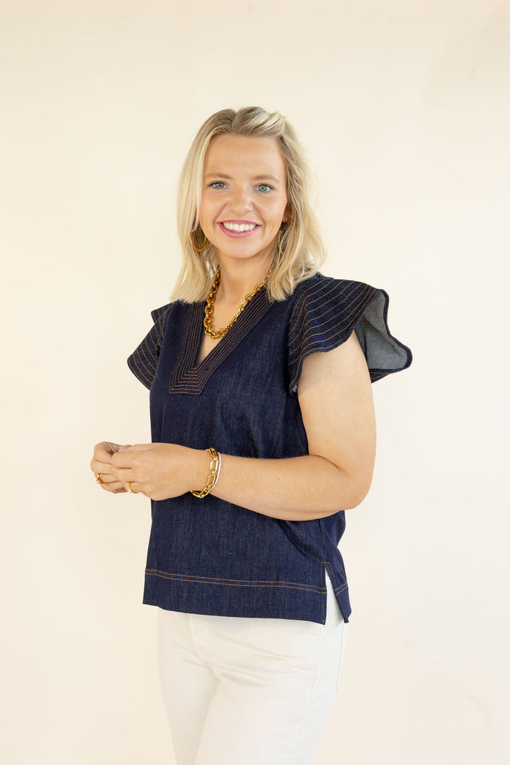 Three-quarter view of woman wearing THML denim blue V-neck top with layered flutter sleeves and contrast stitching styled with white pants
