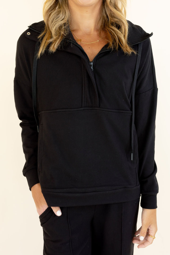 Close-up of black half-zip hooded pullover showing zipper neckline, drawstring hood, long sleeves, and relaxed silhouette