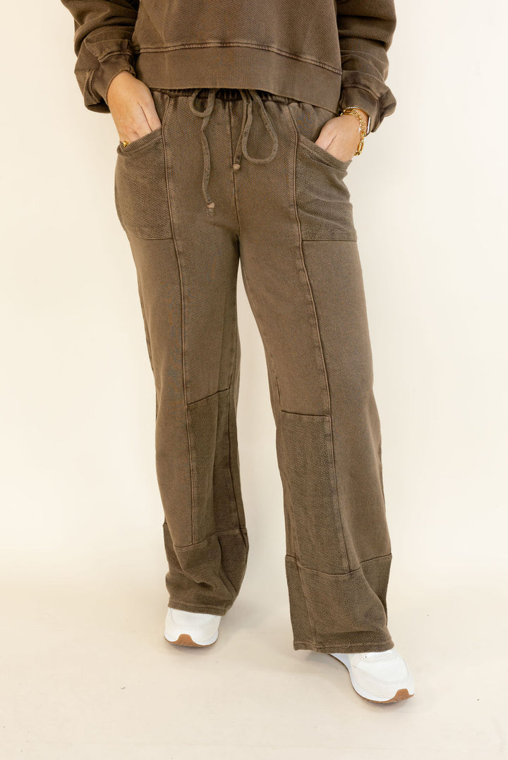 A person wearing loose, wide-leg brown drawstring pants with large front patch pockets, paired with a matching brown sweatshirt and white sneakers, standing against a plain light beige studio background.
