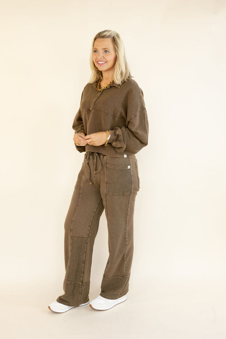 A woman modeling a matching brown two-piece lounge set consisting of a long-sleeve button-up top and wide-leg pants, standing against a plain beige studio background.