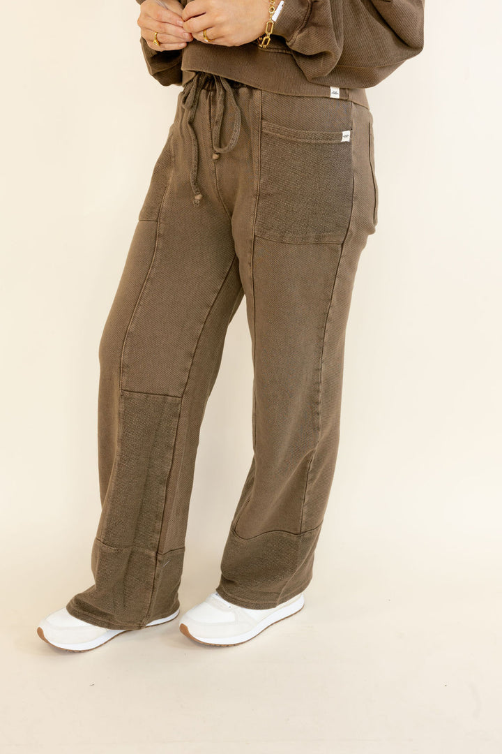 A person wearing loose, wide-leg brown pants with a drawstring waist, large patch pockets, and cuffed hems, paired with white sneakers against a plain light beige studio background.
