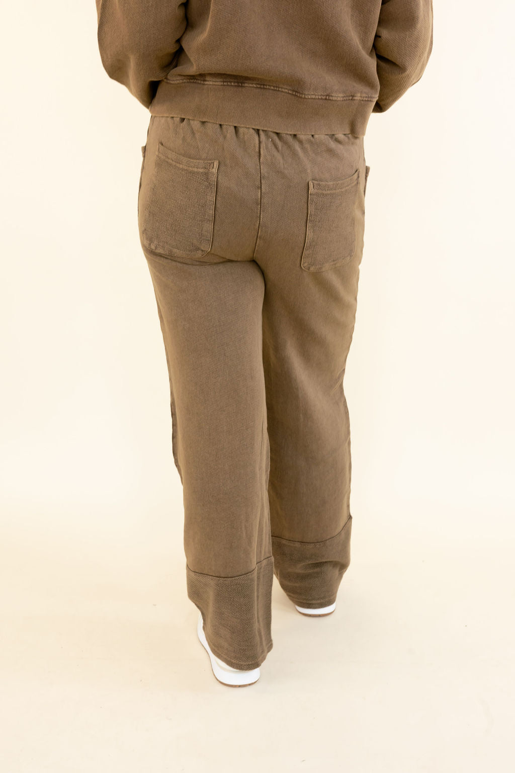 Rear view of a person wearing brown wide-leg pants with a relaxed fit, cuffed hems, and back pockets, paired with a matching brown sweatshirt and white sneakers, against a plain cream background.