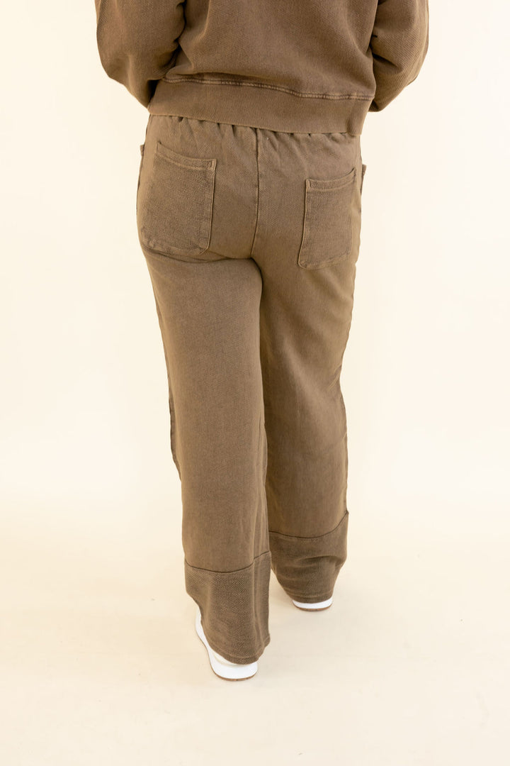Rear view of a person wearing brown wide-leg pants with a relaxed fit, cuffed hems, and back pockets, paired with a matching brown sweatshirt and white sneakers, against a plain cream background.