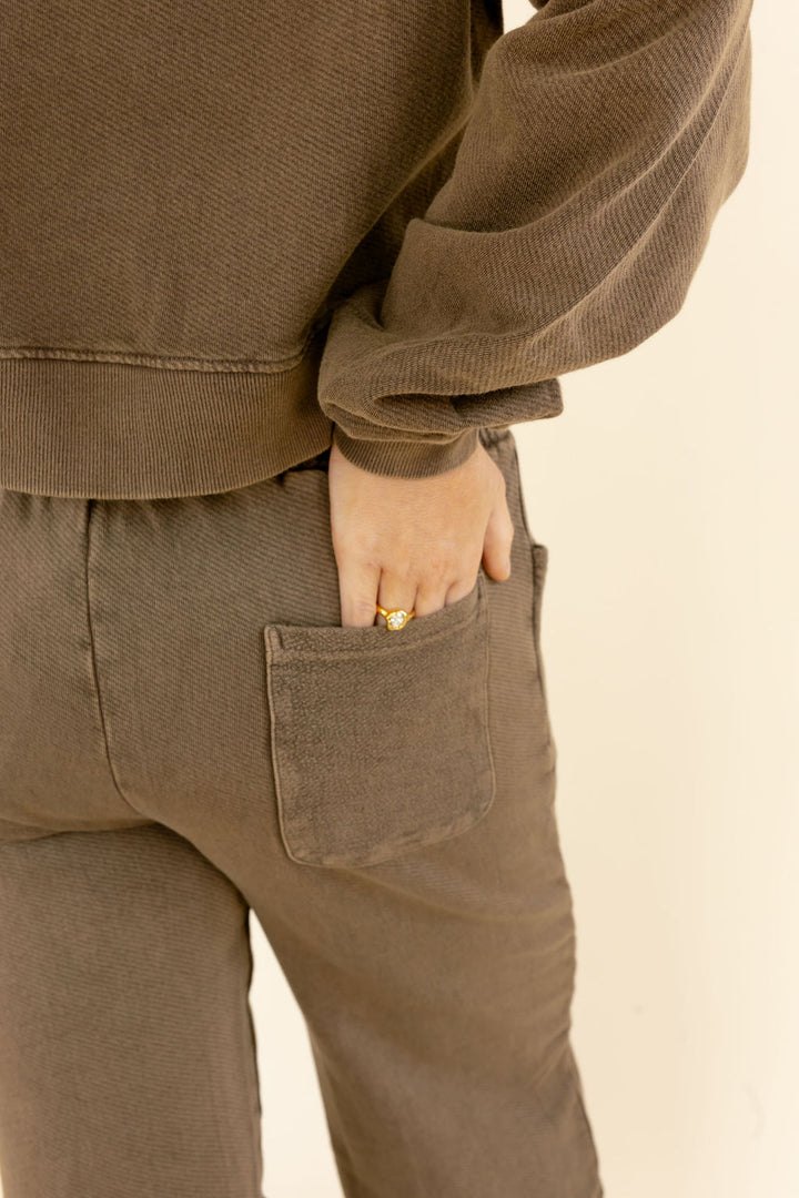 Close-up of a person wearing a matching brown corduroy sweatshirt and pants set, with the hand tucked into the back pocket of the pants.