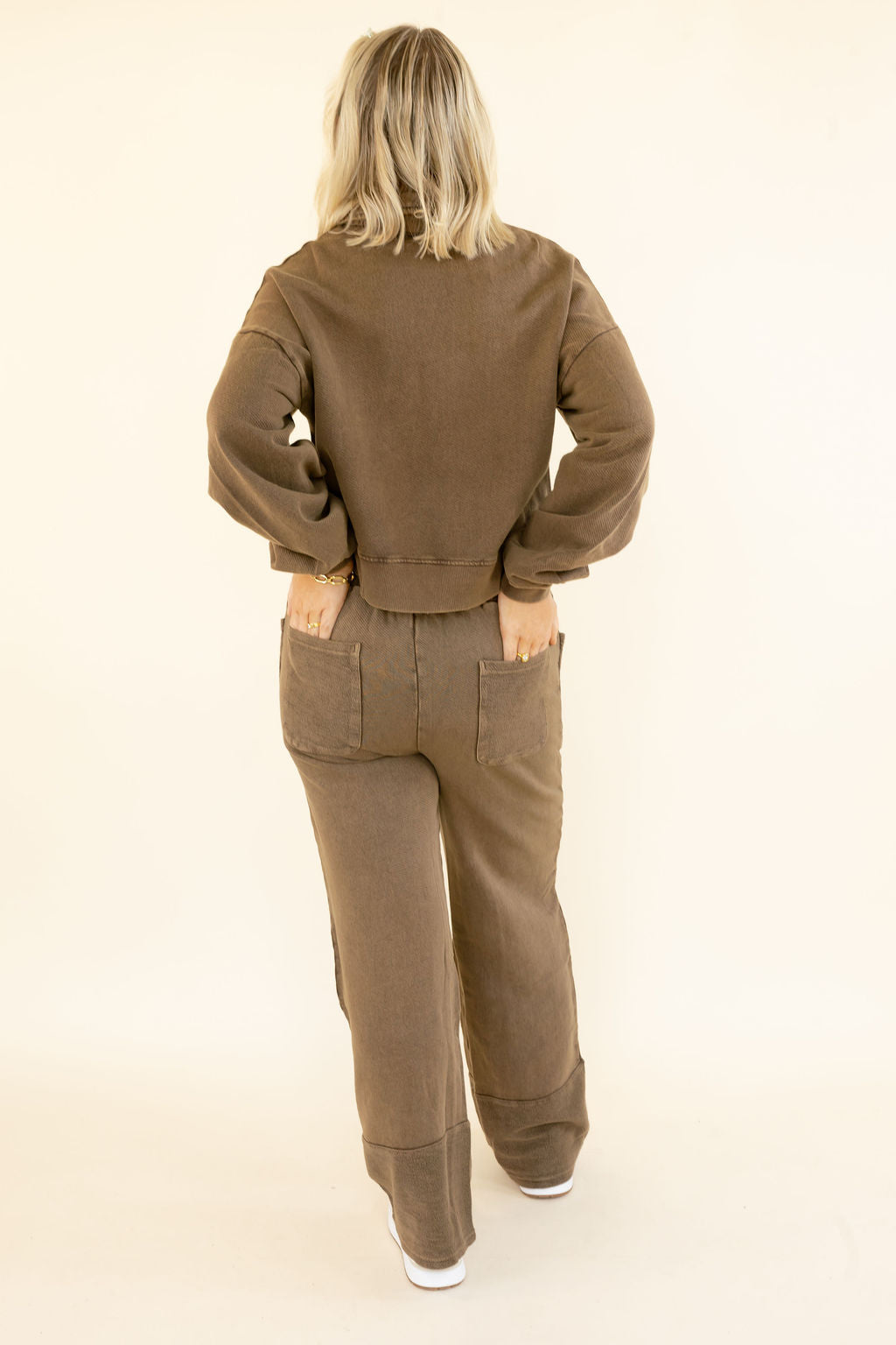 A woman viewed from behind wearing a matching brown sweatshirt and wide-leg pants set, standing against a plain cream studio background.