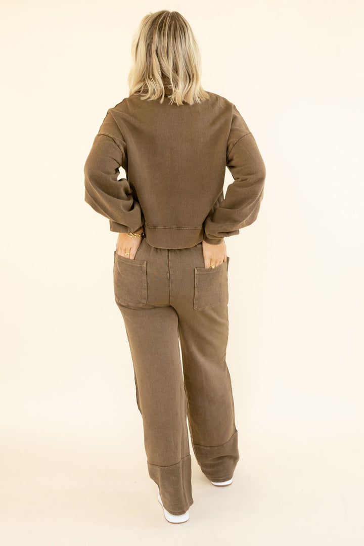 A woman viewed from behind wearing a matching brown sweatshirt and wide-leg pants set, standing against a plain cream studio background.