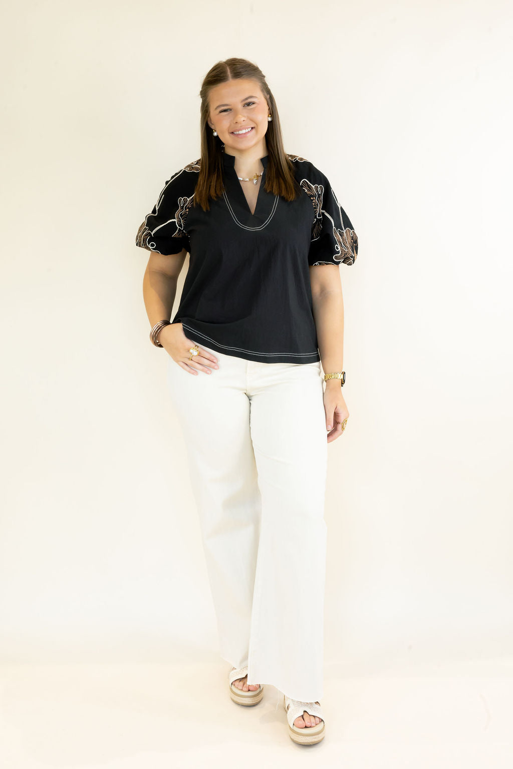 A woman modeling a black short-sleeve top with floral patterned puff sleeves and white wide-leg pants, standing against a plain off-white studio background.