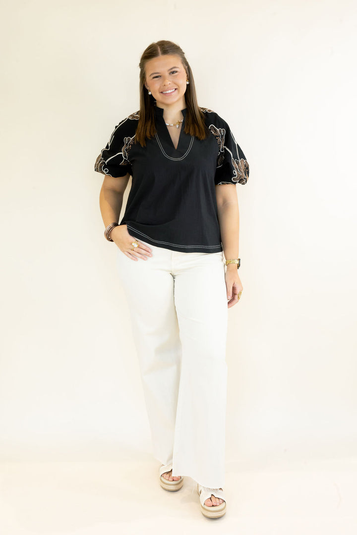 A woman modeling a black short-sleeve top with floral patterned puff sleeves and white wide-leg pants, standing against a plain off-white studio background.