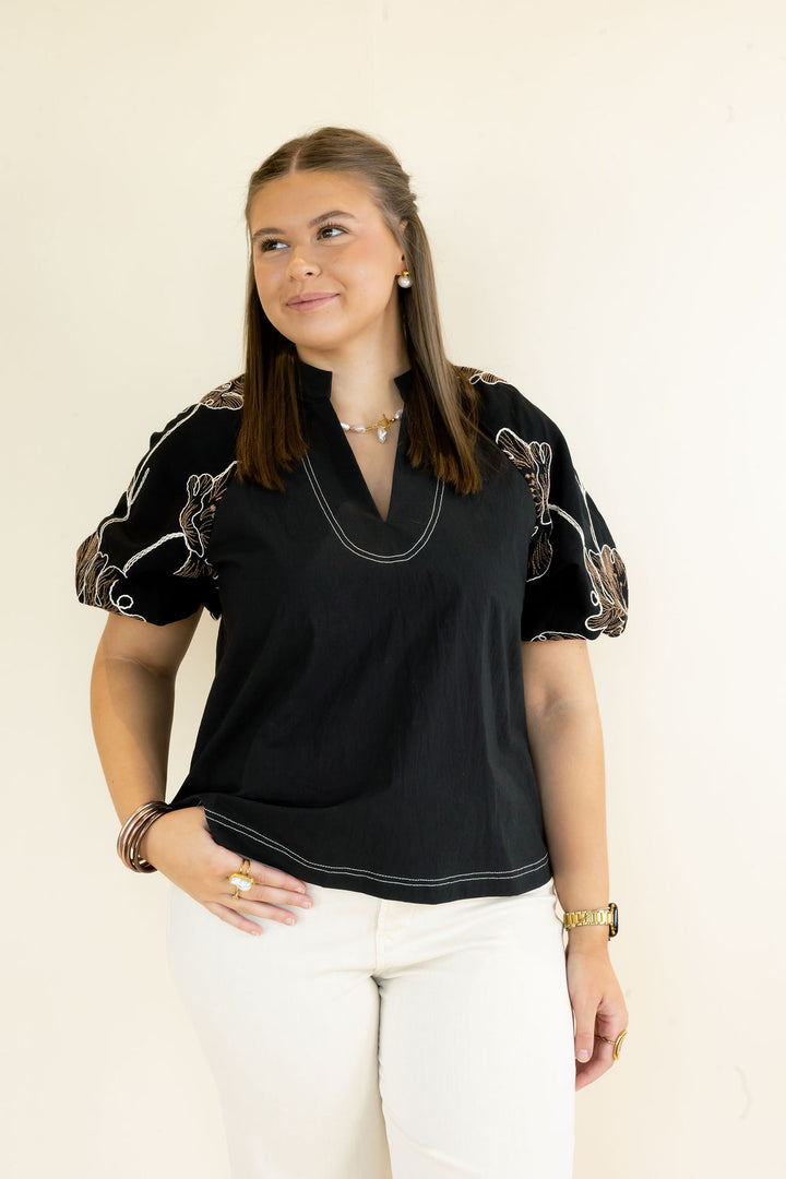 A woman modeling a black short-sleeve blouse with white contrast stitching and gold-toned embroidery on the sleeves, paired with white pants, against a plain off-white background.