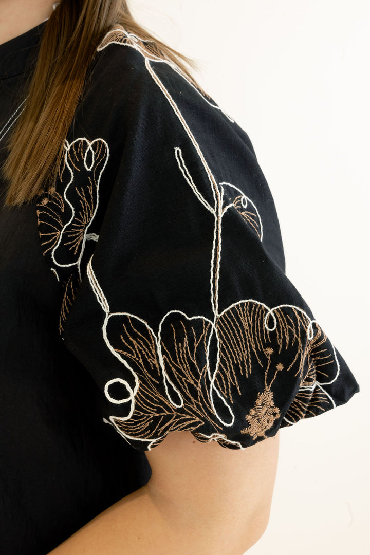 Close-up of a person wearing a black short-sleeve top with large white and gold floral embroidery on the sleeve, against a plain light background.