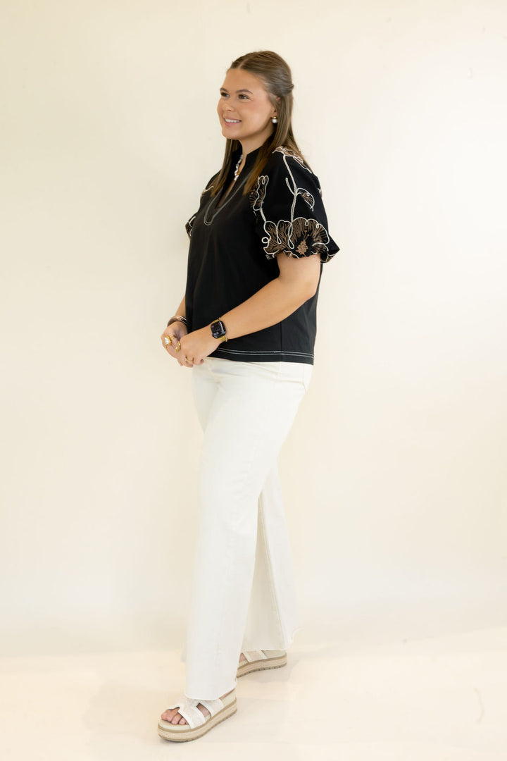 A woman modeling a black short-sleeve top with white embroidered details on the sleeves, paired with white pants and white platform sandals, standing against a plain off-white studio background.