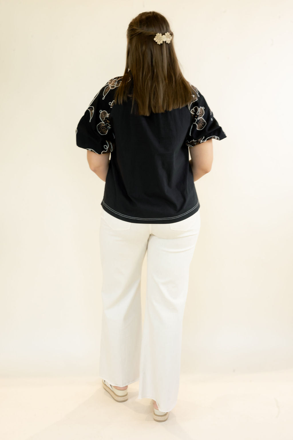 A woman viewed from behind wearing a black short-sleeve top with floral embroidery on the sleeves and white wide-leg pants, standing against a plain off-white studio background.