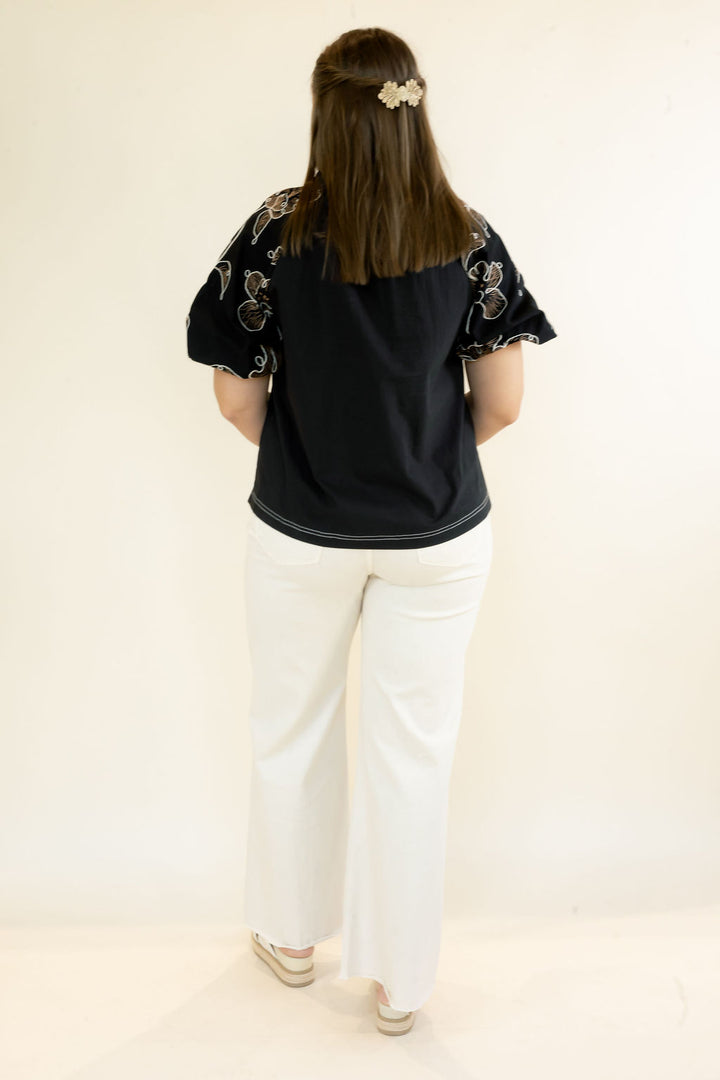 A woman viewed from behind wearing a black short-sleeve top with floral embroidery on the sleeves and white wide-leg pants, standing against a plain off-white studio background.