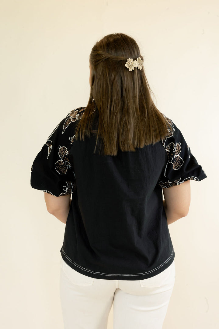 A woman viewed from behind wearing a black short-sleeve blouse with white embroidered details on the shoulders and white pants, standing against a plain off-white background.