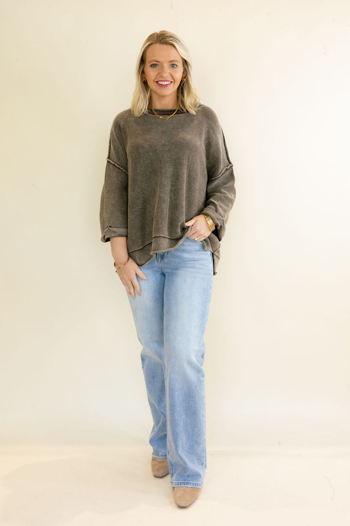 A woman modeling a washed, oversized dark brown knit sweater with cutout details on the sleeves, paired with light-wash blue jeans and beige ankle boots, standing against a plain off-white studio background.