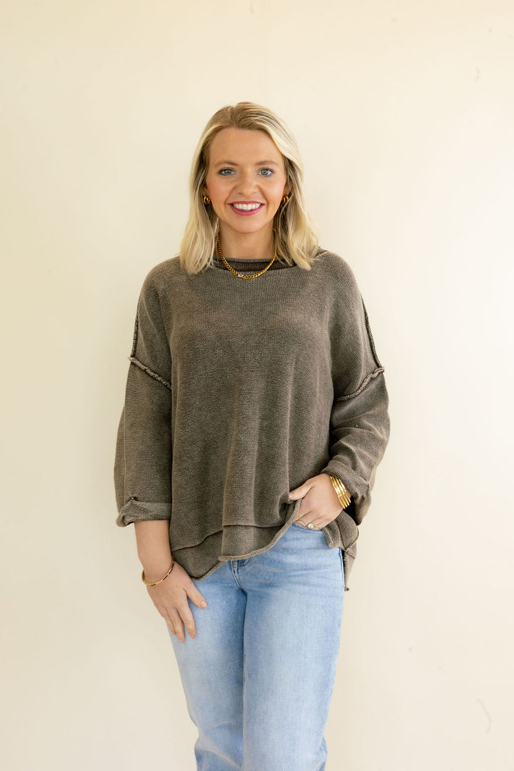 A smiling blonde woman wearing a washed, oversized dark brown long-sleeve knit top and light blue jeans, standing against a plain off-white wall.